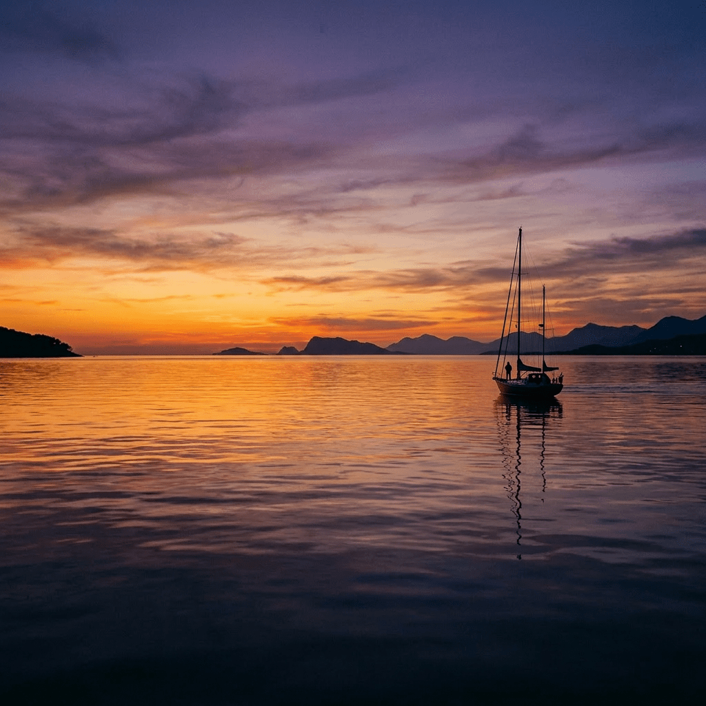 Sailboat silhouetted against orange and purple sunset over calm water with mountains in background